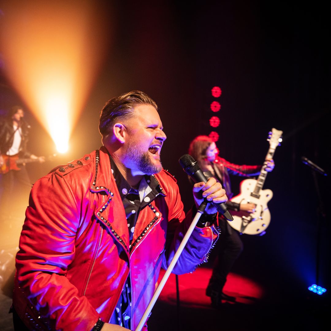 Paradise found production shot-Man singing into mictophone wearing a red leather jacket on stage.